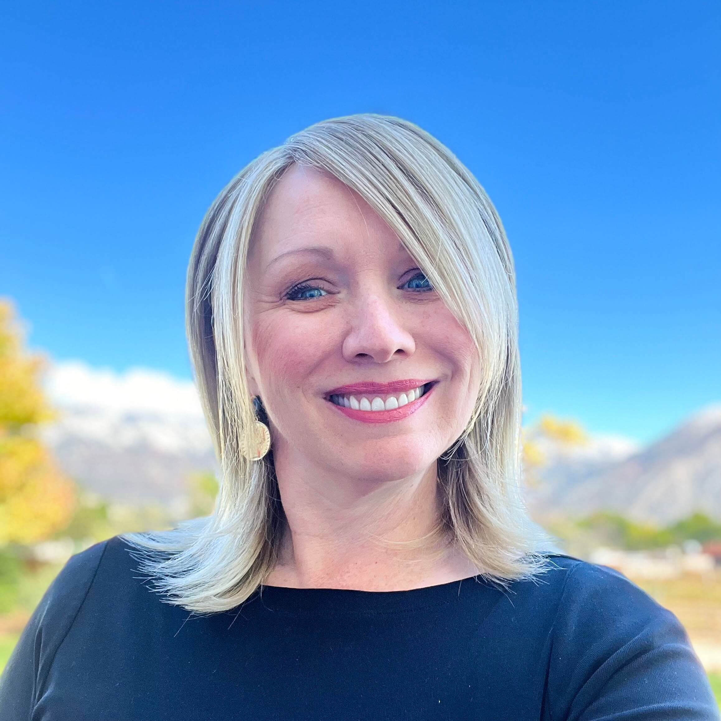 A woman with blonde shoulder-length hair wearing a black top smiles outdoors with mountains, blue sky, and trees in the background.