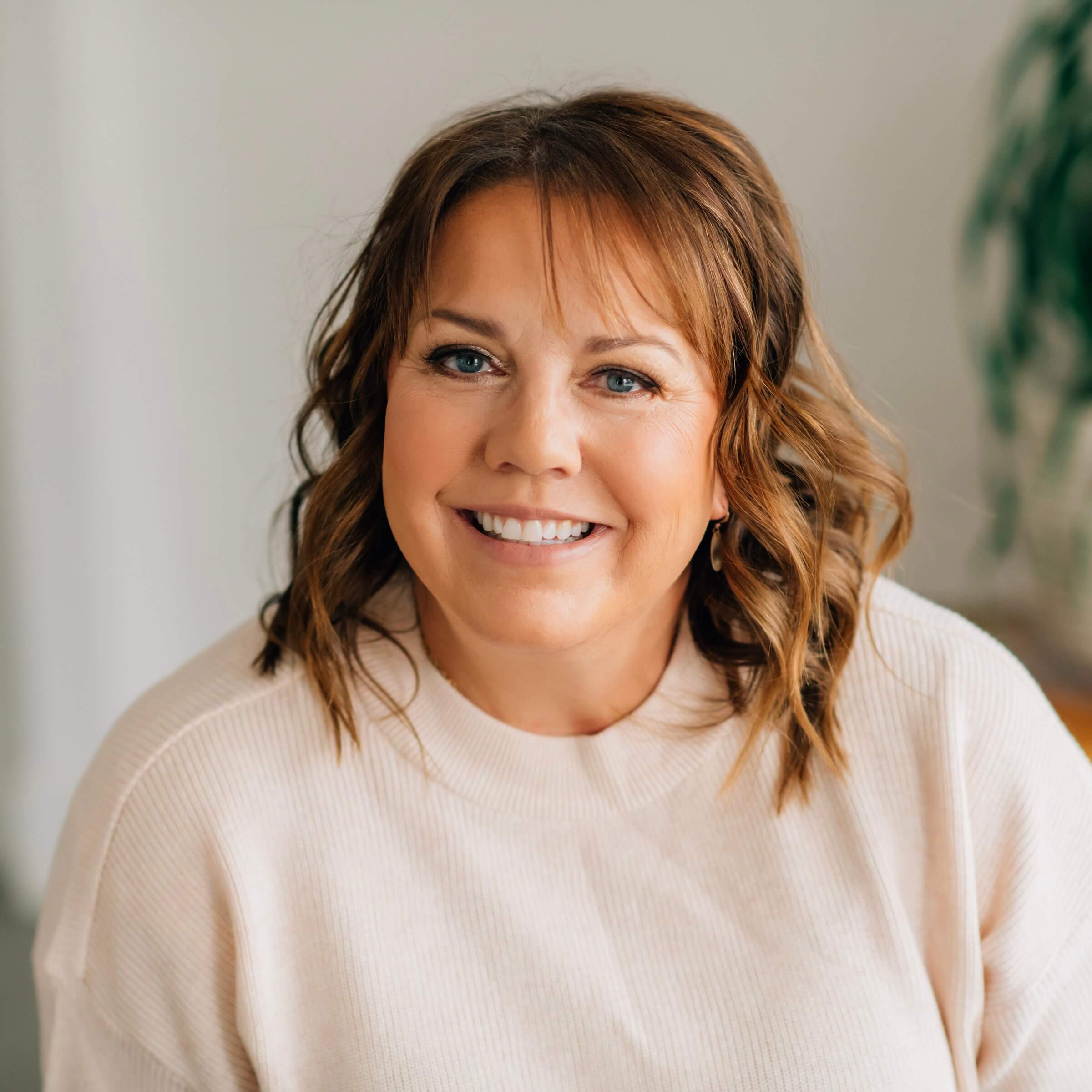 A woman with wavy brown hair wearing a light beige sweater smiles at the camera in a softly lit indoor setting.