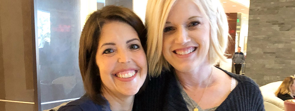 Two women standing close together indoors, smiling at the camera.
