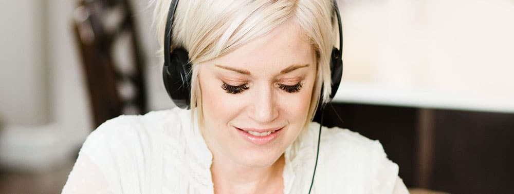 A woman with blonde hair wearing black headphones is looking down and smiling slightly. She is indoors.