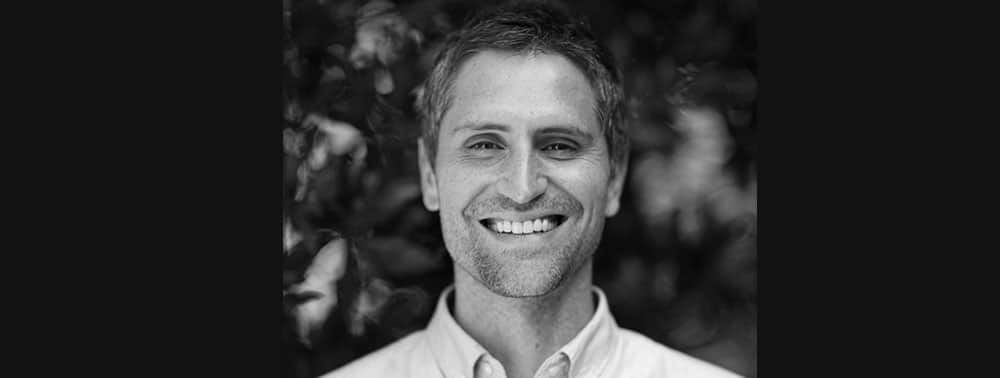 A man with short hair and light stubble is smiling at the camera in a black and white photo with a blurred natural background.