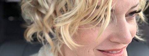 Close-up of a woman with curly blonde hair smiling, showing part of her face and hair in focus.