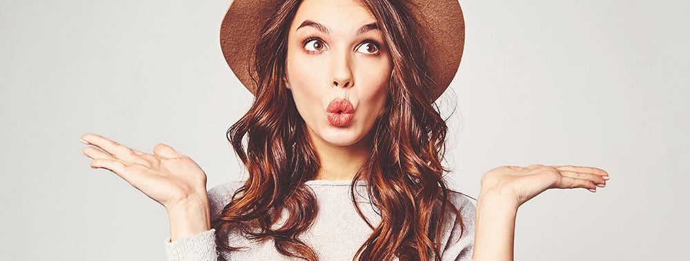A woman with long brown hair wearing a brown hat and light sweater holds her hands up and makes a surprised or puckered expression.