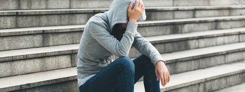 A person in a gray hoodie and jeans sits on outdoor steps with their head down, holding their head with one hand.