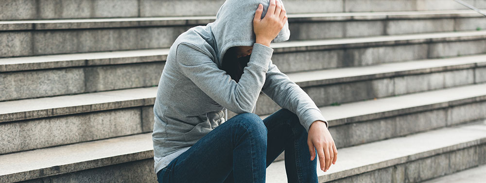 A person in a gray hoodie and jeans sits on outdoor steps with their head down, holding their head with one hand.