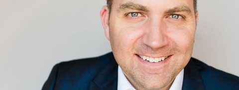 Man in a dark suit smiling at the camera against a plain light background.