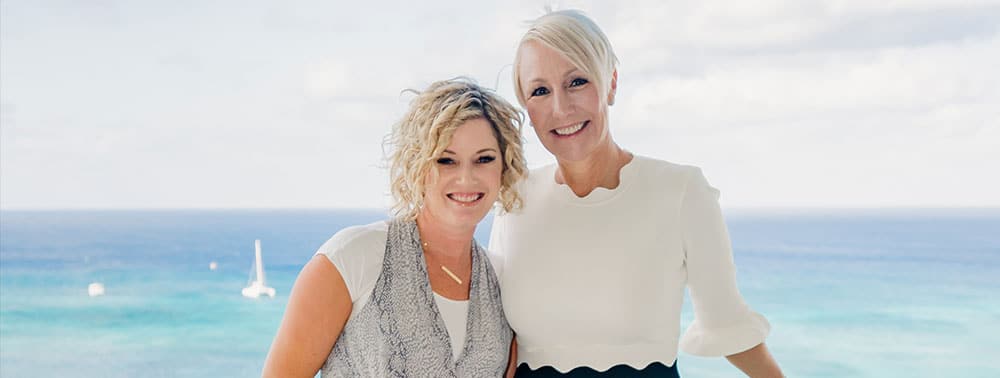 Two women standing together in front of a blue ocean and sky, smiling at the camera.