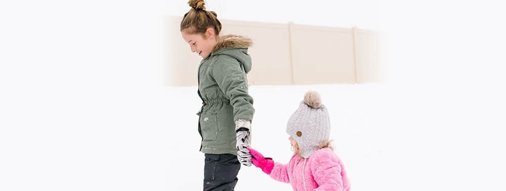 An older child in a green winter coat holds hands with a younger child in a pink coat and gray hat, walking together in the snow.