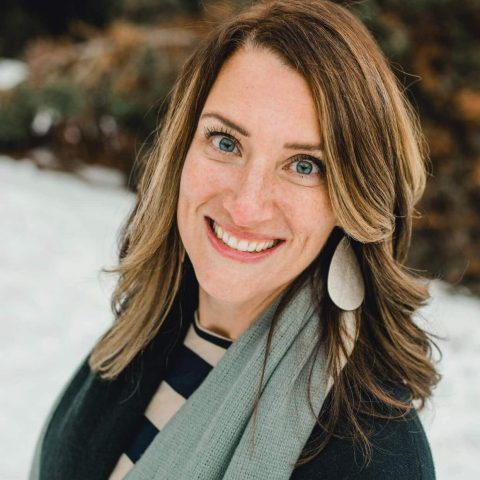 A woman with light brown hair and blue eyes smiles at the camera, wearing a striped shirt, light scarf, and large oval earrings, with a snowy background.