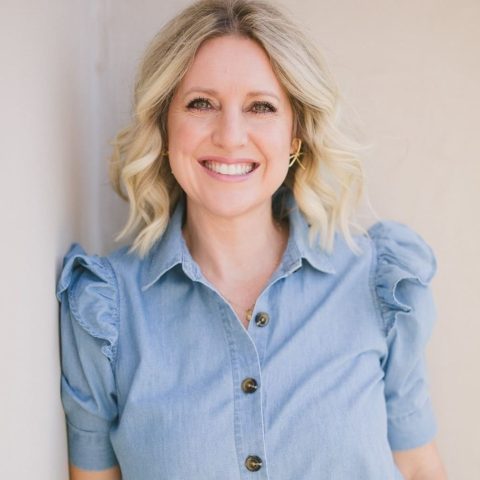 A woman with blonde, wavy hair wears a light blue, short-sleeve, button-up shirt with ruffled shoulders and smiles at the camera.