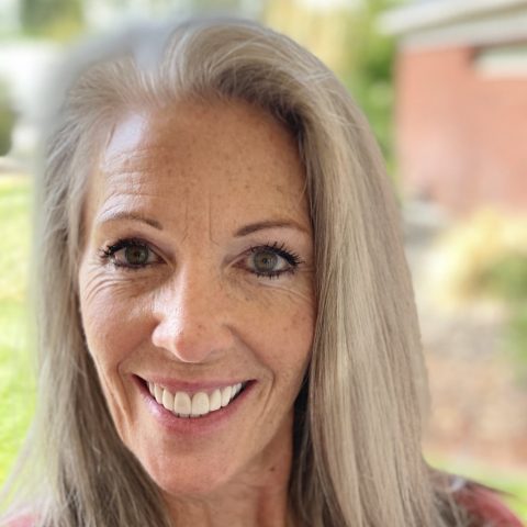 A woman with long gray hair smiles at the camera outdoors, with a blurred background of greenery and a brick building.