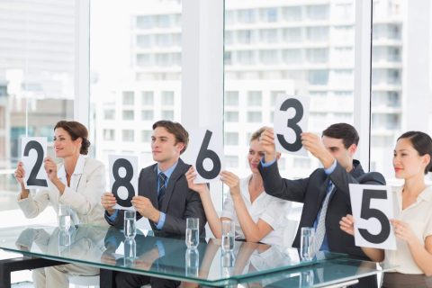 Five people in business attire sit at a glass table, each holding up a number card: 2, 8, 6, 3, and 5, in an office with large windows in the background.