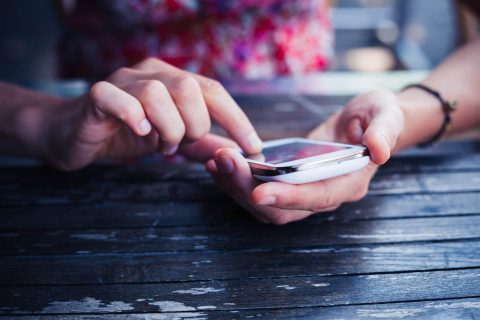 Close-up of a person using a white smartphone at a wooden table, tapping the screen with one hand while holding the phone with the other.
