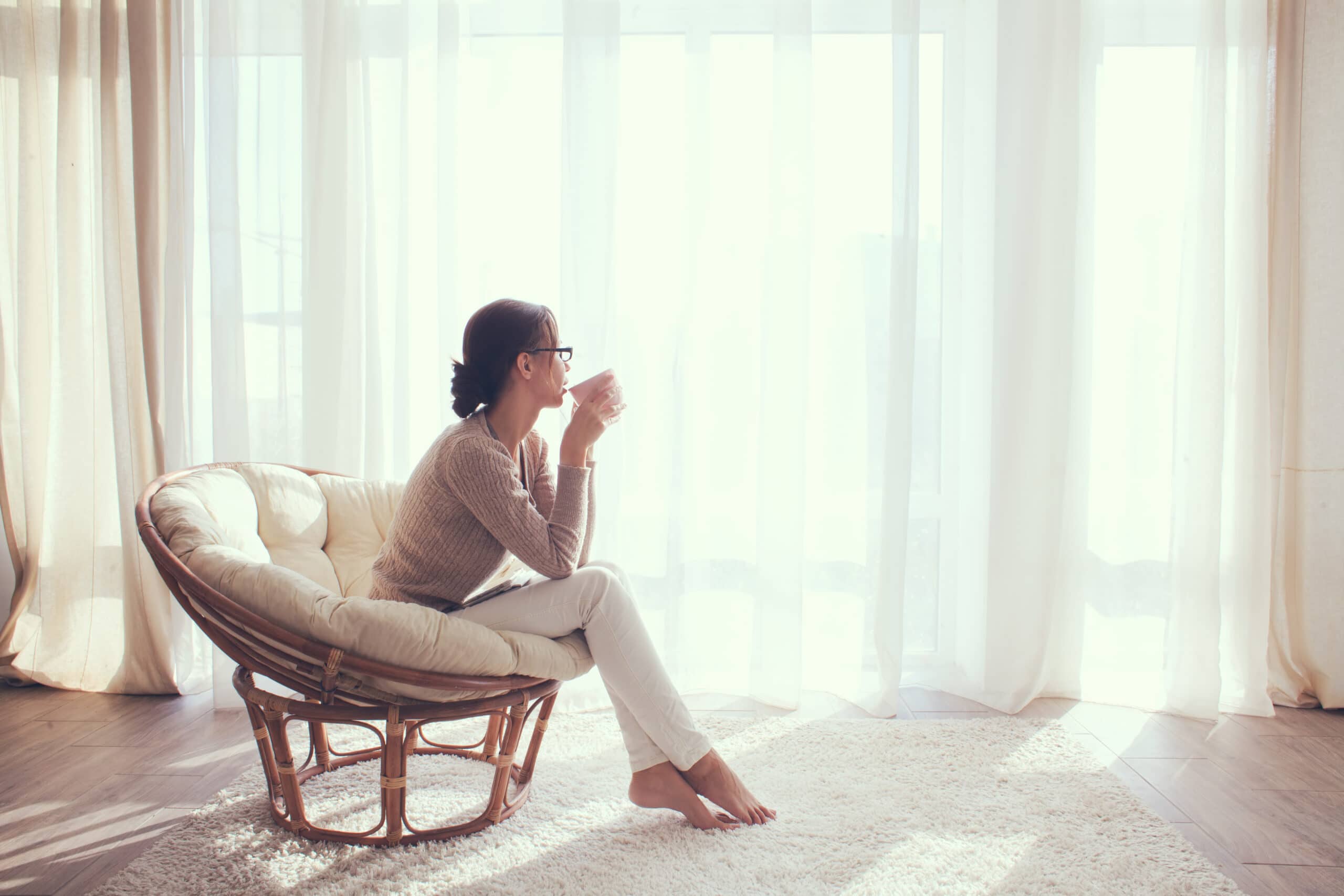 Woman sitting in a cushioned chair by a large window, holding a mug and looking outside, with soft natural light filtering through white curtains.
