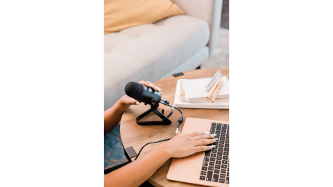 LDS Life Coach using a laptop and holding a microphone at a desk with notebooks and pencils, seated beside a beige sofa.