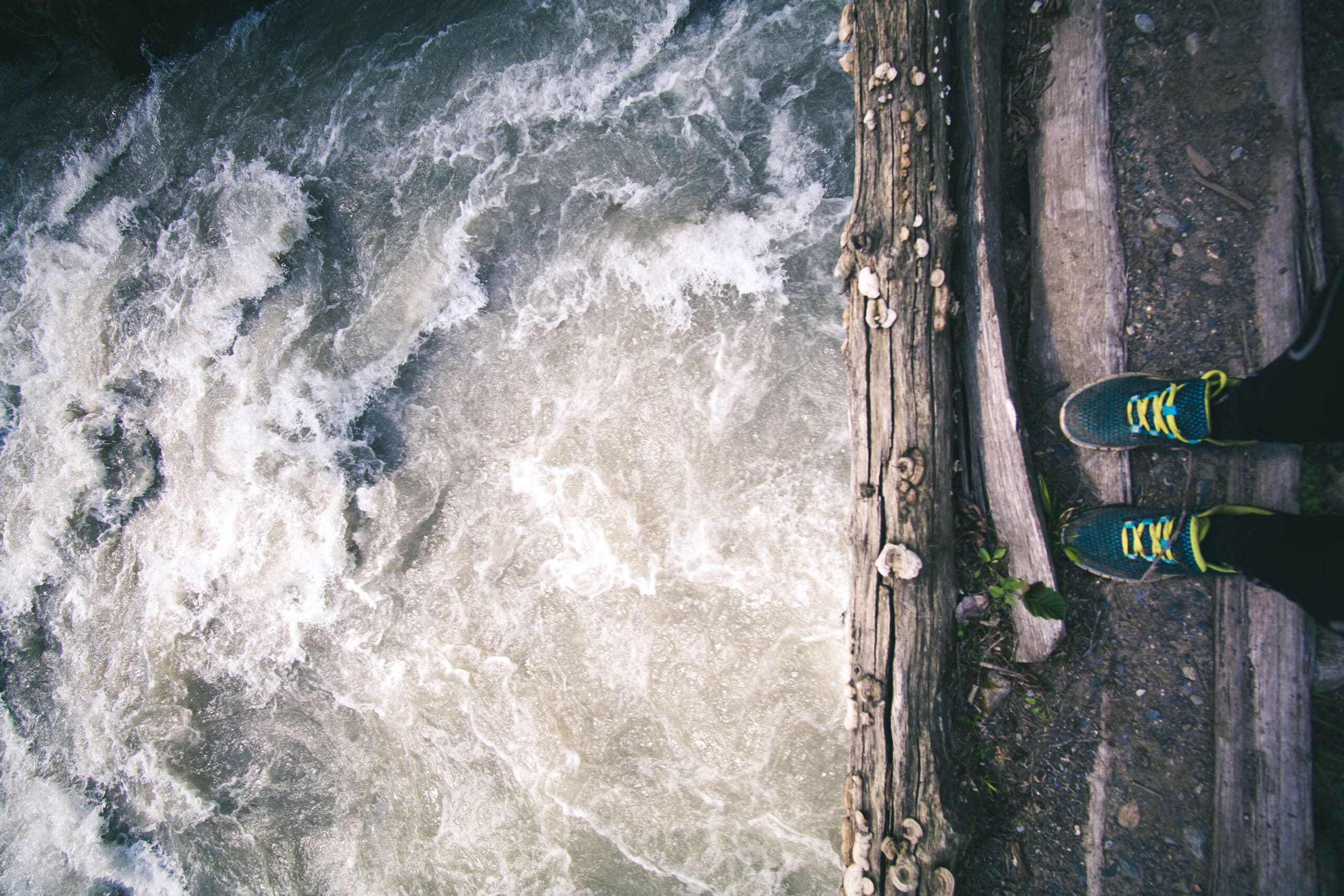 A person wearing blue sneakers stands on wooden planks at the edge of a river with fast-moving water below.