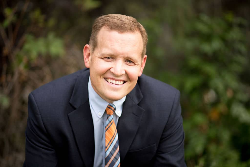 A man in a dark suit, light blue shirt, and striped orange tie smiles at the camera with greenery in the background.