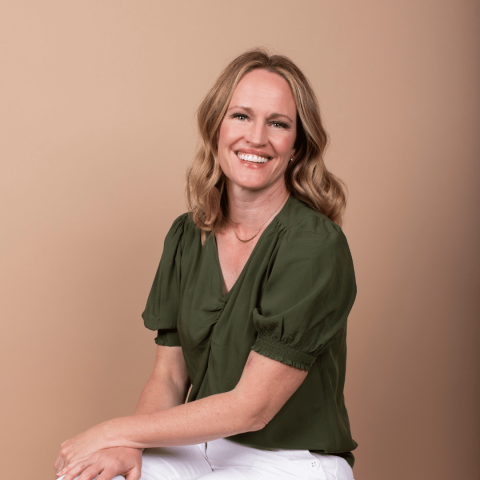 A woman with shoulder-length blonde hair, wearing a green blouse and white pants, sits and smiles against a plain beige background.