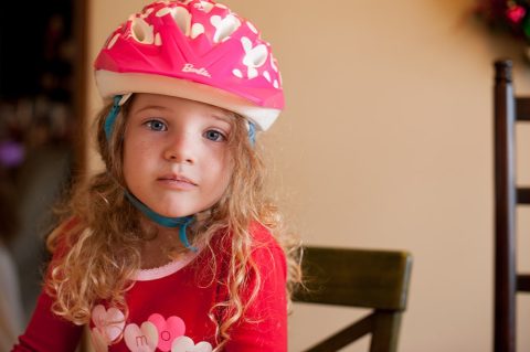Young child with long curly hair wears a pink helmet and a red shirt, sitting indoors on a wooden chair, looking at the camera.