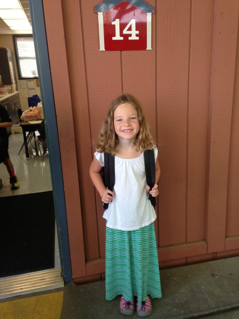 A young girl with a backpack stands and smiles in front of a classroom door marked with a red sign displaying the number 14.