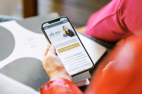 A person holds a smartphone displaying a website for Jody Moore Coaching, highlighting her role as an LDS Life Coach with a photo of a woman and details about free coaching classes.