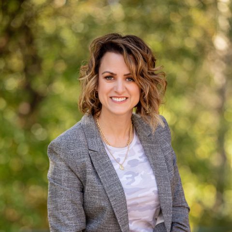 Woman with wavy, shoulder-length hair wearing a gray blazer and white patterned shirt, smiling outdoors with a blurred green background.