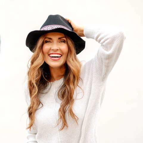 Woman with long wavy hair smiles, wearing a black hat with a patterned band and a white sweater against a plain light background.