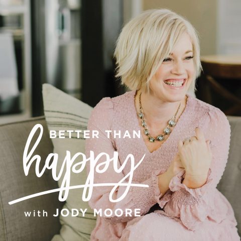 A smiling woman with short blonde hair sits on a couch wearing a light pink dress. Text reads: "Better Than Happy with Jody Moore.