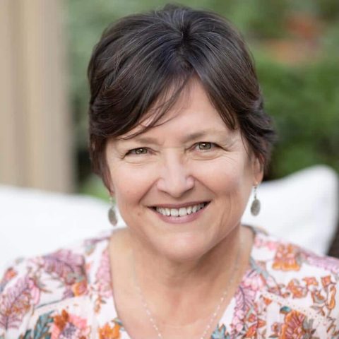 A middle-aged woman with short brown hair, wearing a floral top and earrings, smiles at the camera outdoors.