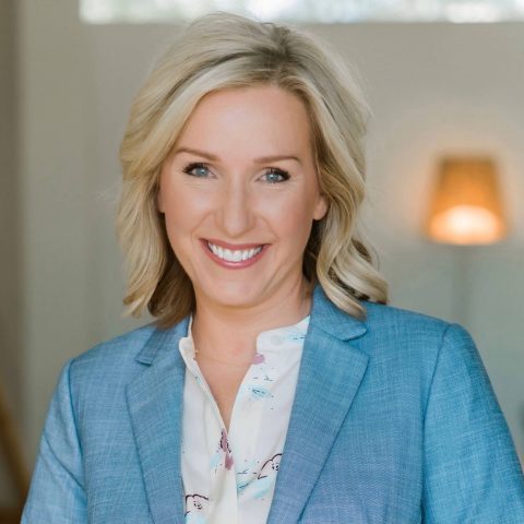 A woman with blonde hair in a blue blazer and patterned shirt smiles at the camera, standing indoors with a blurred lamp in the background.