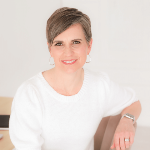 A woman with short brown hair, wearing a white sweater and hoop earrings, sits and smiles at the camera in a bright room.