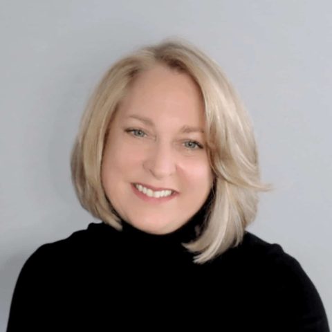 Woman with blonde, shoulder-length hair wearing a black turtleneck, smiling in front of a plain light gray background.