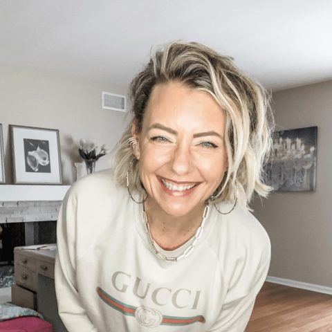 A woman with short blonde hair smiles at the camera, wearing a cream Gucci sweatshirt, chain necklace, and hoop earrings in a light-colored living room.