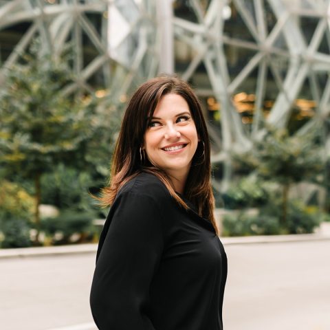 A woman with brown hair wearing a black top smiles while standing outdoors in front of a modern glass building.