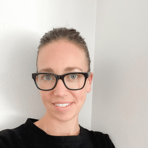 A person with brown hair tied back, wearing black glasses and a black top, is standing in front of a white wall and smiling at the camera.