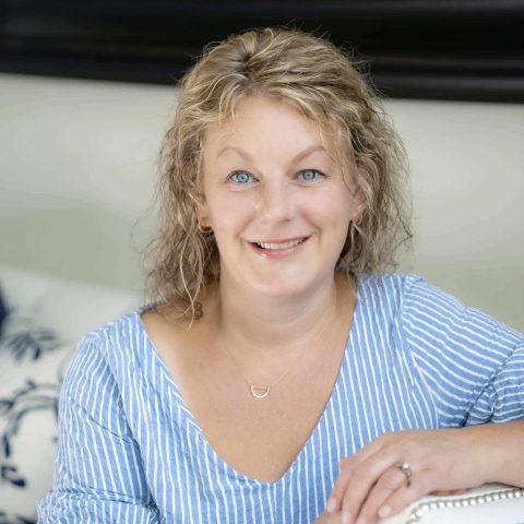 A woman with curly blonde hair and blue eyes, wearing a blue and white striped blouse, sits indoors and smiles at the camera.
