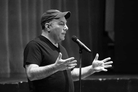 A man in a black t-shirt and cap speaks into a microphone on stage, gesturing with both hands. The image is in black and white.