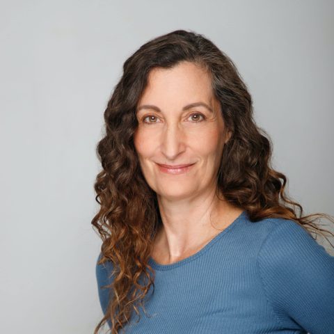 A woman with long curly brown hair and a blue shirt smiles slightly while facing the camera against a plain light background.
