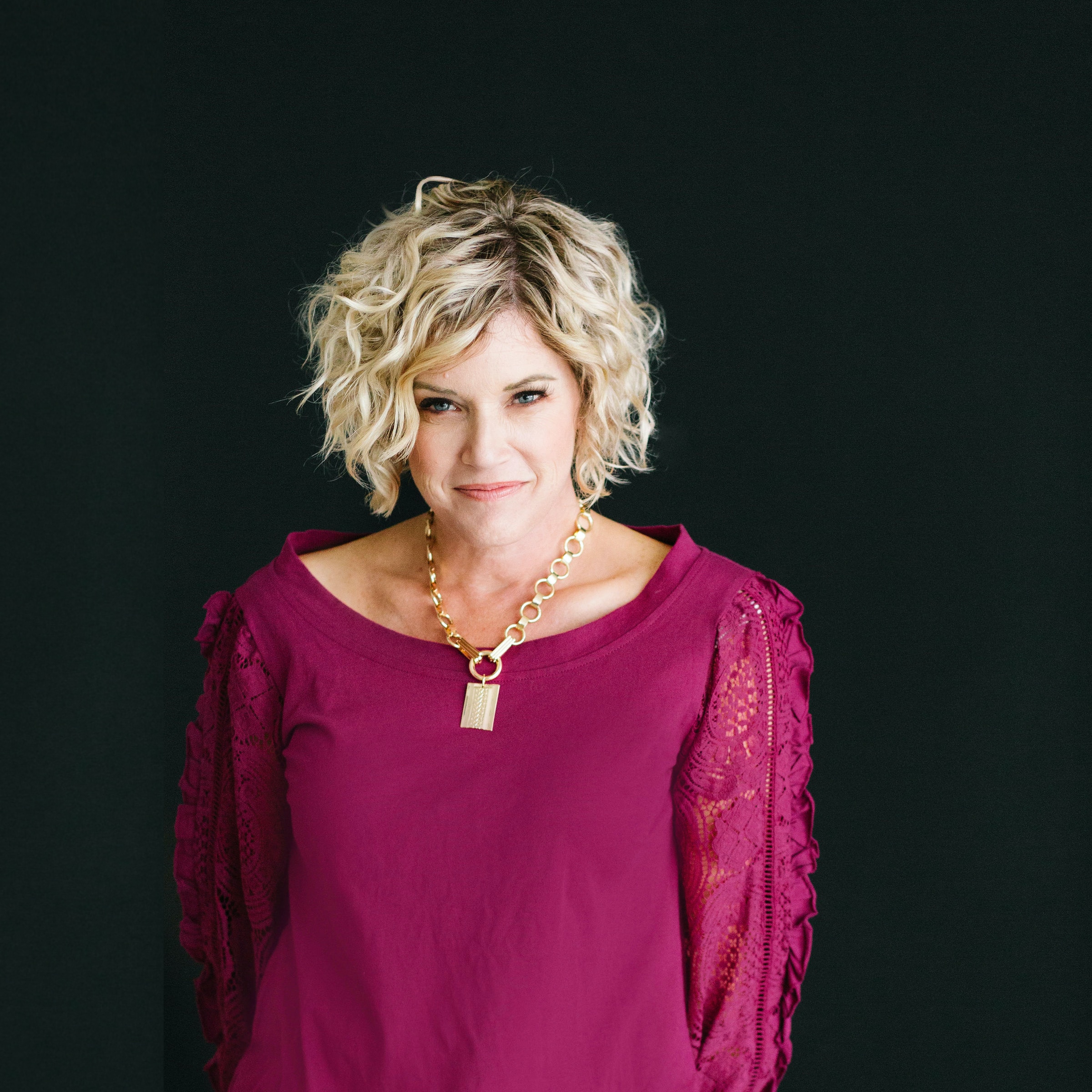 A woman with short curly blonde hair wears a magenta top with lace sleeves and a chunky gold necklace, standing against a plain black background.