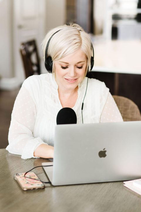 A woman wearing headphones speaks into a microphone while working on a laptop at a table. A smartphone and notebook are placed nearby.