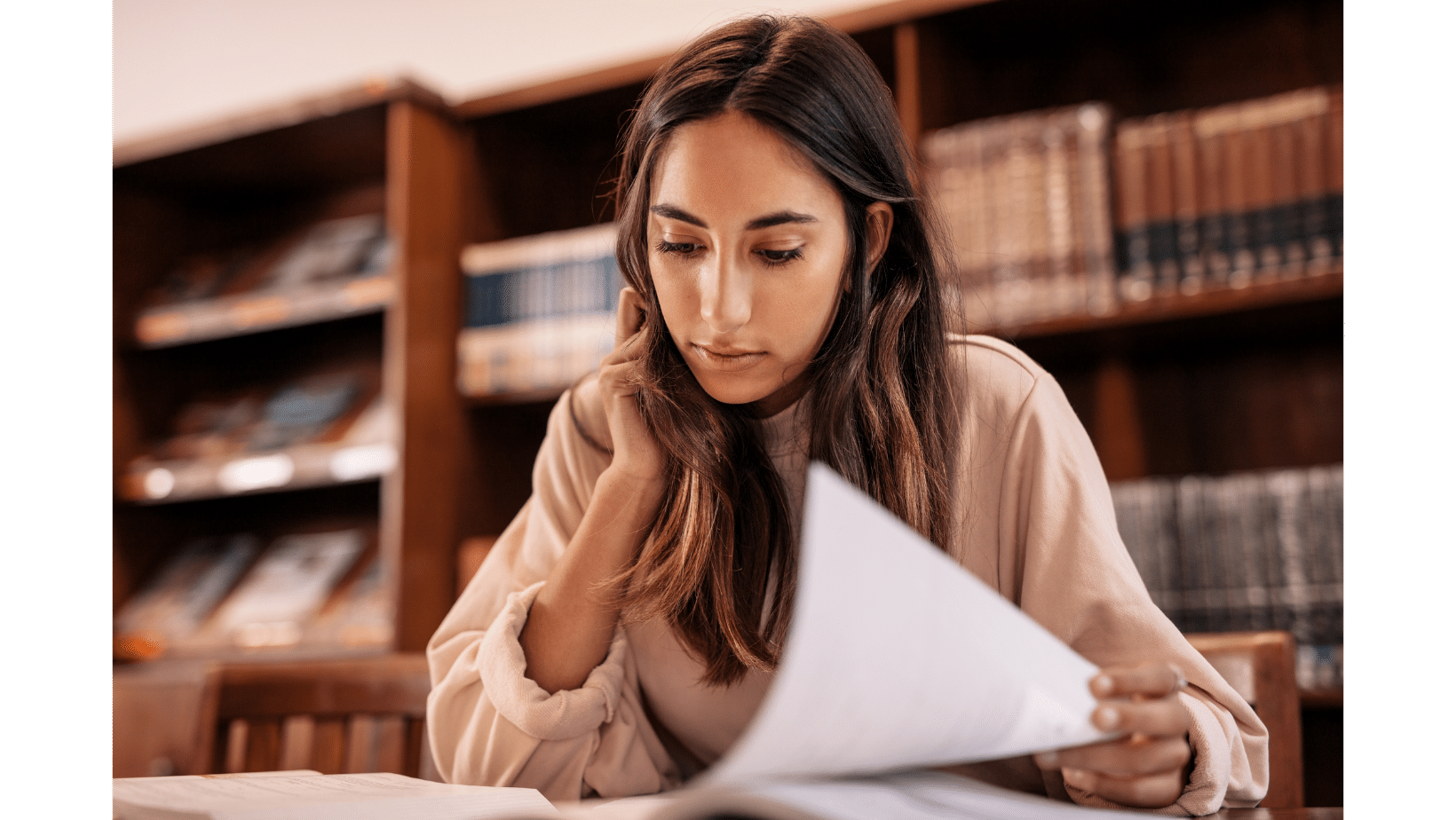 A woman sits at a table in a library, reading and holding papers, navigating her faith and questions in the LDS faith, with bookshelves in the background.