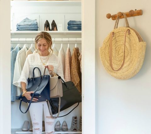 A woman stands in a closet holding two handbags, with clothes, shoes, and accessories neatly organized around her; a straw bag hangs on a hook outside the closet.