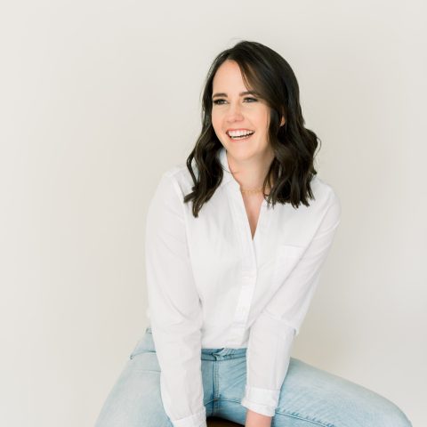 A woman with shoulder-length dark hair, wearing a white shirt and light blue jeans, sits and smiles against a plain light background.