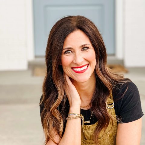 A woman with long brown hair and a bright red lipstick smiles at the camera, resting her hand on her neck. She wears a black shirt and tan overalls, sitting in front of a blurred background.