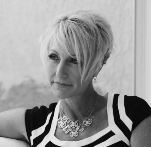 Black and white photo of a woman with short, light hair, wearing a patterned top and a statement necklace, looking slightly to the side with a neutral expression.