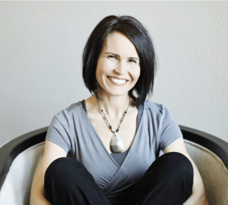 A woman with straight, dark hair sits in a chair, smiling. She wears a gray shirt and a large pendant necklace, with her arms resting on her knees.