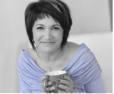 Woman with short dark hair wearing a light-colored top, smiling and holding a mug with both hands while seated indoors.