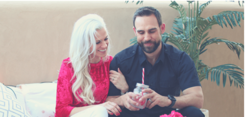A woman in a pink top and a man in a navy shirt sit together on a couch, smiling and sharing a drink with a striped straw. A potted plant is in the background.
