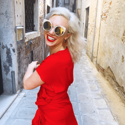 Woman with blonde hair wearing large round sunglasses and a bright red dress smiles broadly while walking down a narrow stone alleyway.