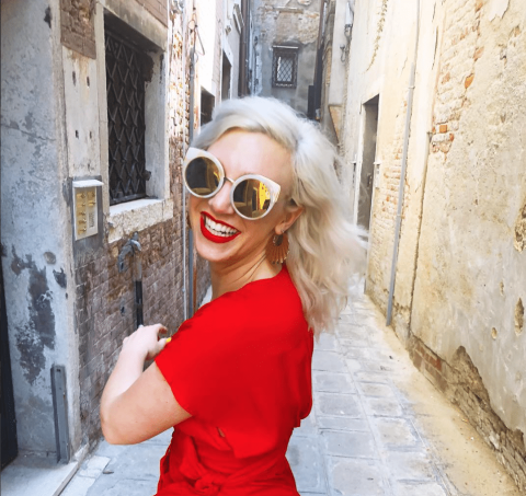 Woman with platinum blonde hair, wearing round sunglasses and a red dress, smiles while standing in a narrow, sunlit alleyway with stone walls and pavement.
